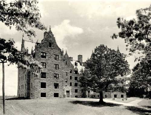 Historic stone building with gothic architecture, surrounded by trees and a grassy area under a cloudy sky.