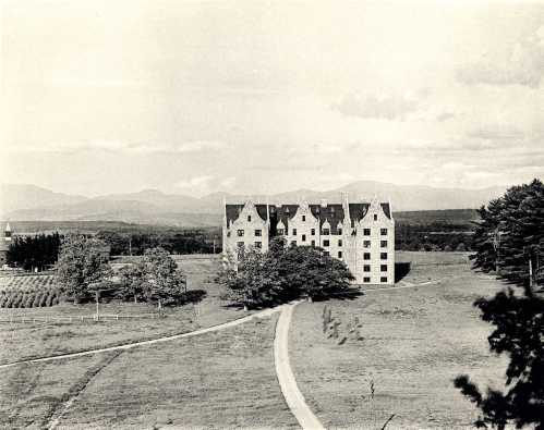 A historic building surrounded by trees and fields, with mountains in the background under a cloudy sky.