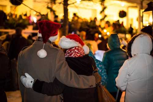 A couple in Santa hats embraces, surrounded by a festive crowd at a holiday event.