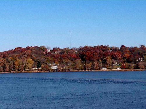 A serene lake view with a hillside covered in autumn foliage and a tall radio tower in the background.