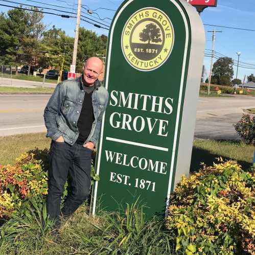 A man stands next to a welcome sign for Smiths Grove, Kentucky, established in 1871, surrounded by greenery.