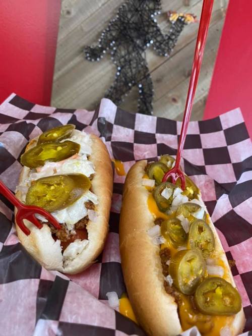 Two hot dogs in a checkered paper basket, topped with jalapeños, onions, and served with red plastic forks.