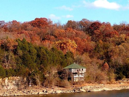 A rustic house sits by a river, surrounded by vibrant autumn foliage and a clear blue sky.