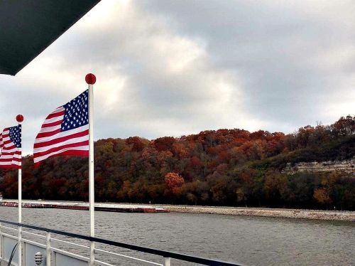 Two American flags flutter on a boat, with colorful autumn trees lining the riverbank under a cloudy sky.