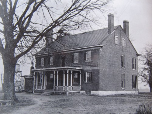 Historic two-story brick house with a porch, surrounded by trees, in a black-and-white photograph.