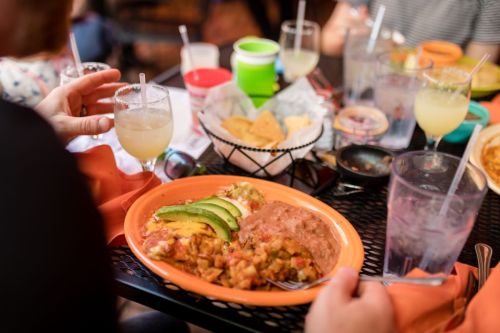 A colorful plate of food with avocado, beans, and chips, alongside a drink, on a table with other dishes.
