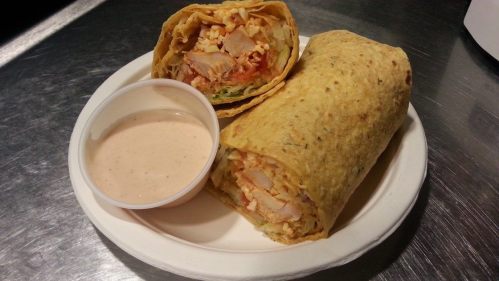 Two wraps filled with chicken and vegetables on a plate, accompanied by a small cup of dipping sauce.
