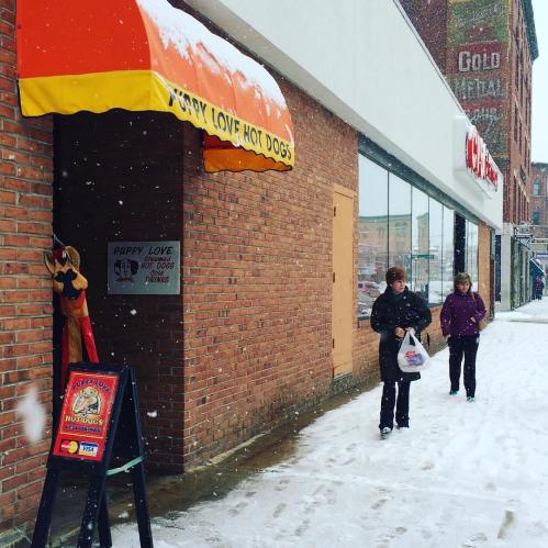 A snowy street scene with two people walking past a hot dog stand, "Puppy Love Hot Dogs," under a bright orange awning.