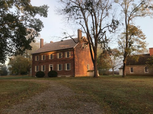 A historic two-story wooden house surrounded by trees and a grassy area, with a gravel path leading to it.