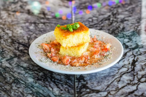 A plate of cornbread topped with green onions, surrounded by shrimp on a marbled table.