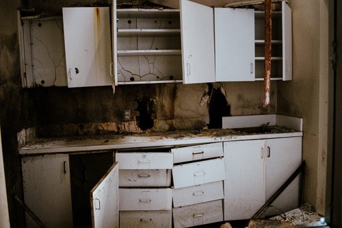An abandoned kitchen with cracked walls, open cabinets, and scattered debris on the counter.