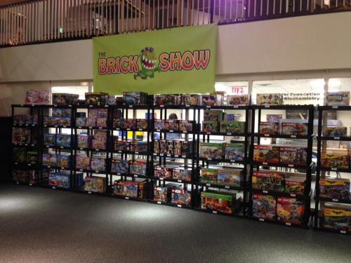 A display of various LEGO sets on shelves, with a banner reading "The Brick Show" in the background.