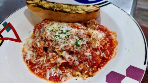 A plate of cheesy lasagna topped with marinara sauce and herbs, served with a slice of garlic bread.