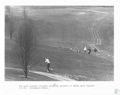 A golfer swings on a hillside at Devou Golf Course, with others walking in the background on a warm day.