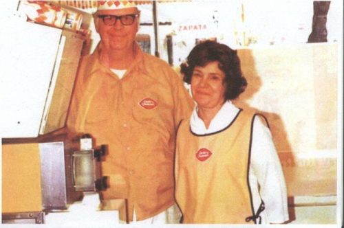 A man and woman in vintage uniforms stand together in a store, both wearing name tags and smiling at the camera.