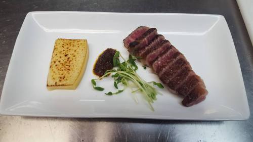 A plated dish featuring sliced steak, a piece of grilled polenta, and garnishes of microgreens and sauce.