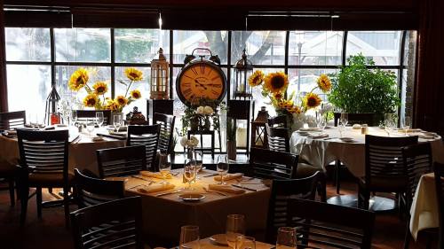 Cozy restaurant interior with tables set for dining, sunflowers, lanterns, and a large clock by a window.