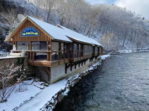 A cozy riverside cabin with a snow-covered roof, surrounded by trees and a flowing river.