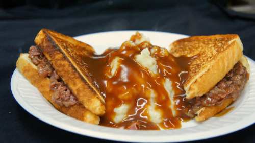 A plate with two toasted sandwiches filled with meat, served alongside mashed potatoes topped with brown gravy.