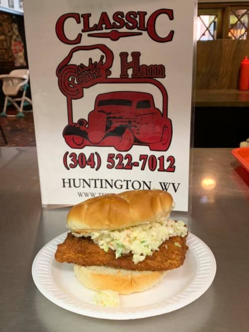 A crispy fried sandwich with coleslaw on a bun, placed on a paper plate in front of a restaurant sign.