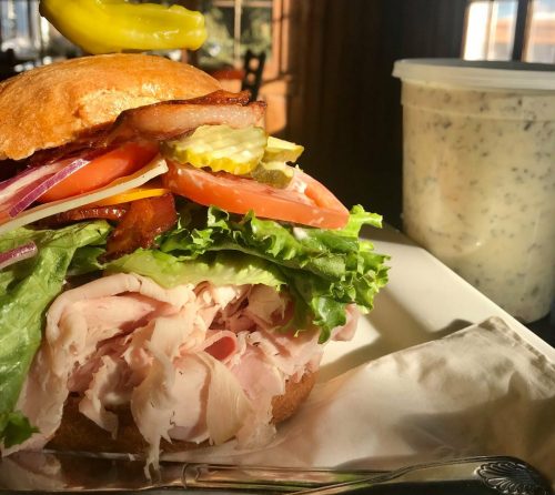 A close-up of a hearty sandwich with turkey, lettuce, tomatoes, pickles, and a pepper on top, with a container of dressing nearby.