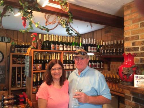 A man and woman smile together in a wine shop, surrounded by shelves of bottles and decorative elements.