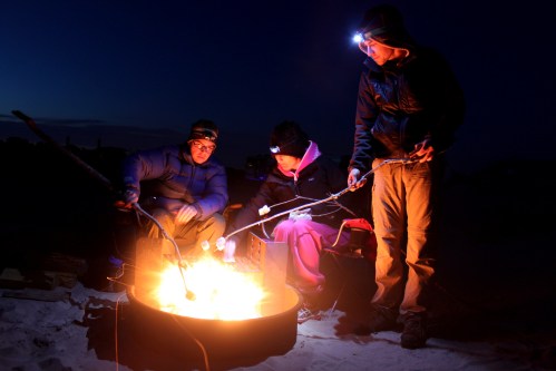 Three people gather around a campfire at night, roasting marshmallows and wearing headlamps.
