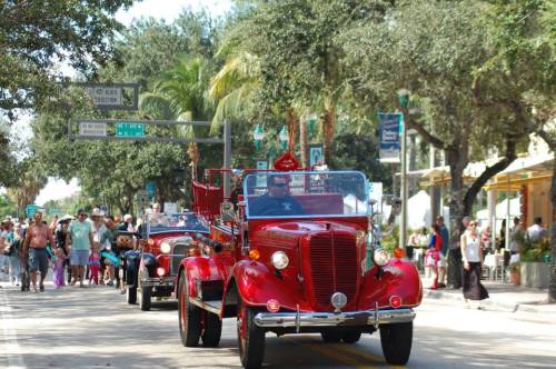A vintage red fire truck leads a parade down a tree-lined street with people watching.
