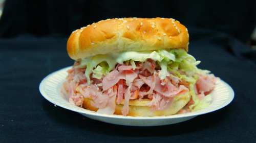 A sesame seed bun sandwich filled with shredded ham, lettuce, and mayonnaise on a paper plate.