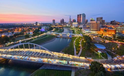 A vibrant city skyline at dusk, with a river, bridges, and illuminated buildings reflecting in the water.