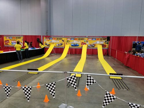 A colorful Pinewood Derby track with yellow ramps, checkered flags, and people preparing for a race event.