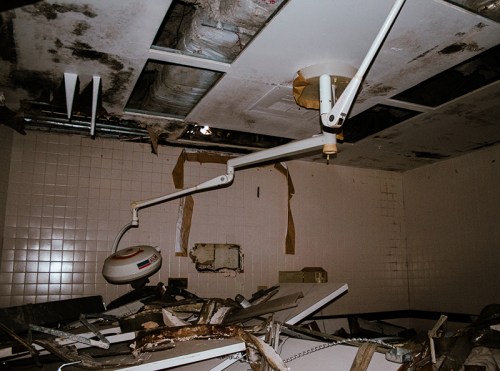 An abandoned room with a broken medical light and debris scattered on the floor, showing signs of decay and neglect.