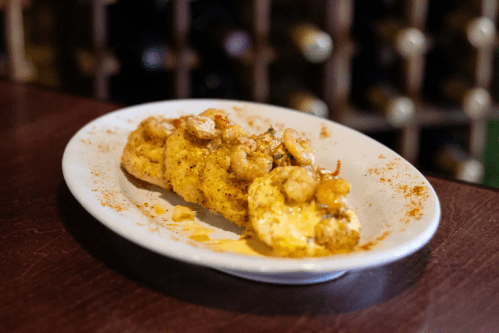 A plate of shrimp and grits, garnished with spices, served on a wooden table with a wine rack in the background.