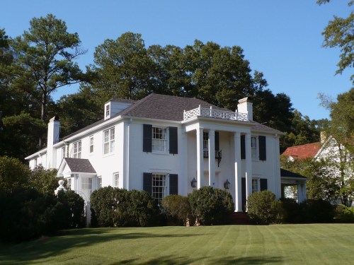 A large white house with black shutters, surrounded by greenery and trees under a clear blue sky.