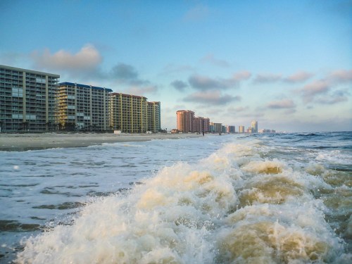 Waves gently roll onto a sandy beach lined with tall buildings under a blue sky with scattered clouds.