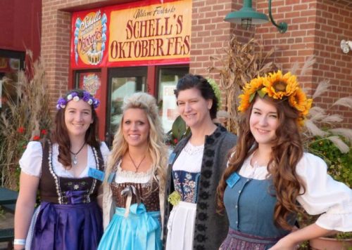 Four women in traditional dresses and flower crowns pose in front of a festive Oktoberfest sign.