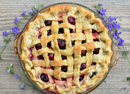 A cherry pie with a lattice crust, surrounded by purple flowers on a wooden surface.