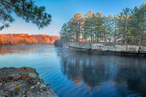 A serene river scene with mist, surrounded by trees and rocky cliffs under a clear blue sky.
