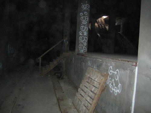 Dimly lit interior of a building with graffiti on the walls and a wooden pallet on the floor near a staircase.