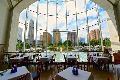 A restaurant interior with tables set for dining, featuring large windows showcasing a city skyline and blue sky.