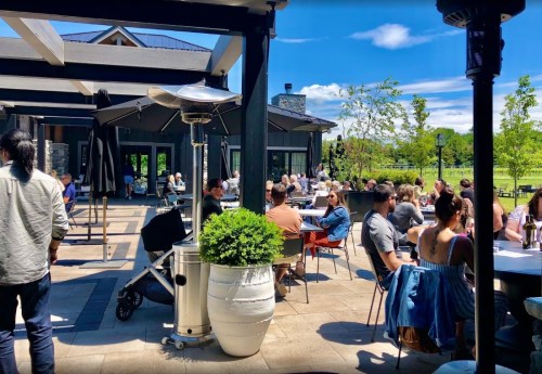 A lively outdoor dining area with people seated at tables, enjoying a sunny day surrounded by greenery.