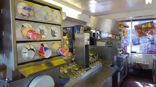 Interior of an ice cream shop with a machine dispensing soft serve and colorful cups stacked in holders.