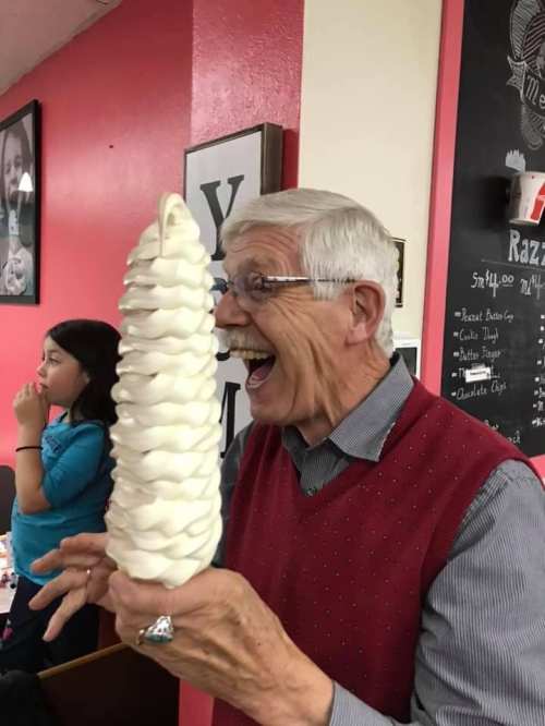 An elderly man joyfully holds a large swirl of soft serve ice cream, smiling widely in a colorful diner setting.