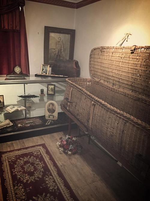 A dimly lit room featuring a wicker coffin, framed pictures, and a display case with various artifacts.