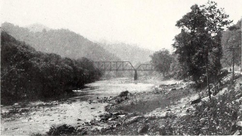 Black and white image of a river with a bridge in the background, surrounded by hills and trees.