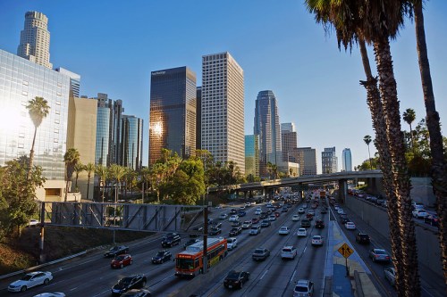 A busy Los Angeles highway with palm trees and skyscrapers under a clear blue sky.