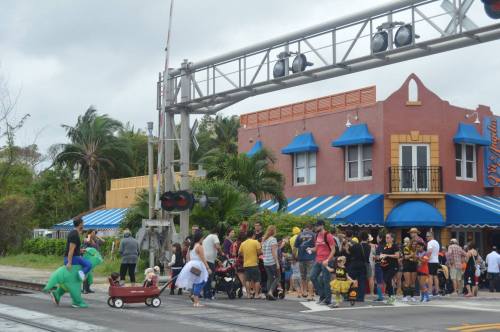 A crowd gathers near a train crossing, with colorful buildings and palm trees in the background. A person in a dinosaur costume is visible.