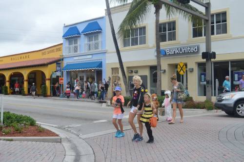 A group of children in costumes crossing the street near shops, with a crowd and palm trees in the background.