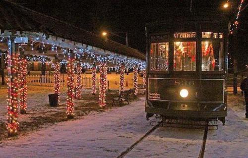 A decorated streetcar on snowy tracks, surrounded by festive lights and a charming station at night.
