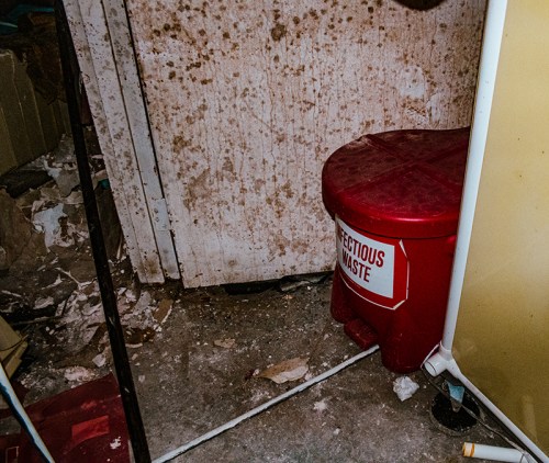 A red waste bin labeled "Biohazard" sits in a cluttered, moldy room with debris on the floor.